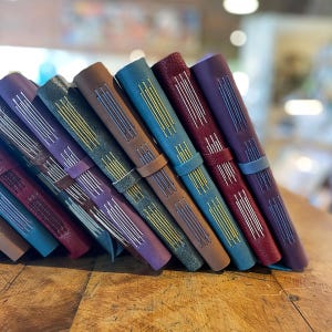 May include: A collection of handmade leather journals in various colors, including teal, brown, and purple. Each journal features hand-stitched binding with contrasting thread and a leather strap closure. The journals are arranged on a wooden surface.