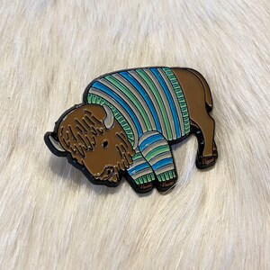 May include: A brown and black enamel pin of a bison wearing a blue, green, and white striped sweater.