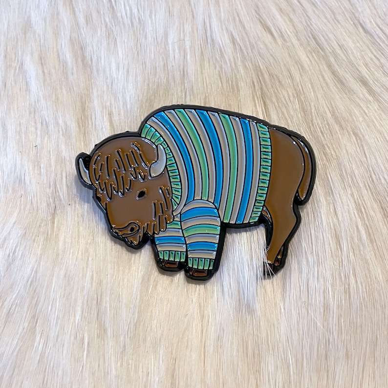 May include: A brown and black enamel pin of a cartoon bison wearing a blue, green, and white striped sweater.