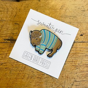 May include: A brown and white enamel pin of a bison wearing a blue and green striped sweater. The pin says "sweaterpin" and "GREEN BIRD PRESS" on the card.