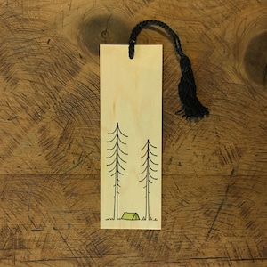 May include: A wooden bookmark with a black tassel. The bookmark features a simple illustration of two pine trees and a green tent.