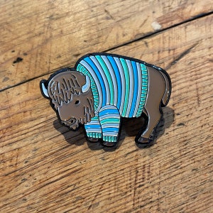 May include: A brown and black enamel pin of a bison wearing a blue and green striped sweater.