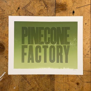 May include: A print with the words "PINECONE FACTORY" in dark green block letters on a light green gradient background. The print is on white paper.