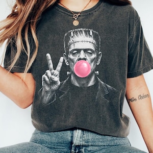 Puede incluir: Una camiseta de manga corta gris carb&oacute;n con un gr&aacute;fico en blanco y negro del monstruo de Frankenstein soplando una burbuja de chicle de color rosa brillante. El monstruo hace el signo de la paz con una mano y viste una chaqueta oscura.