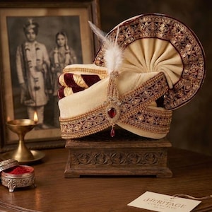 Op de afbeelding: Een sierlijke crèmekleurige tulband met bordeauxrode en gouden versieringen, een juwelen hanger en een witte veer. De tulband wordt getoond op een gesneden houten kist, met een vintage foto in een lijst op de achtergrond.