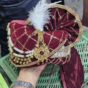 Op de afbeelding: Een bordeauxrode fluwelen tulband met gouden versieringen, een witte veer en parel- en gouden accenten. De tulband heeft een decoratief goud en robijnrood juweel. Het ontwerp is sierlijk en traditioneel.