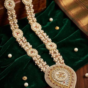 Puede incluir: Un collar dorado adornado con múltiples hilos de pequeñas perlas blancas. El collar presenta elementos decorativos circulares y en forma de lágrima con detalles rojos y blancos. La joya descansa sobre una superficie de terciopelo verde oscuro.