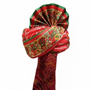 Pode incluir: Um turbante vermelho com um padrão floral e paisley detalhado. O turbante apresenta uma borda verde e dourada e um design decorativo. O tecido é dobrado e enrolado, criando um adorno de cabeça tradicional.