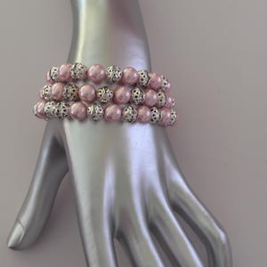 Könnte beinhalten: Dreireihiges Armband mit rosa Perlen und silbernen Filigranakzenten. Das Armband wird auf einer silbernen Hand präsentiert, die das Design und die Farbe hervorhebt. Die Perlen sind ein zartes Rosa, und das Filigran sorgt für einen Hauch von Eleganz.