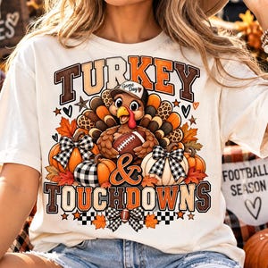 May include: A cream-coloured t-shirt featuring a graphic of a turkey wearing a baseball cap and holding a football. The design includes pumpkins, autumn leaves, and the text "TURKEY & TOUCHDOWNS" in brown and orange block lettering.