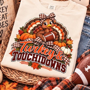 May include: A cream-coloured t-shirt featuring a graphic of a turkey wearing a plaid bow, holding a football, surrounded by pumpkins and autumn leaves, with the text "Turkey & TOUCHDOWNS" in orange and brown lettering on the front.