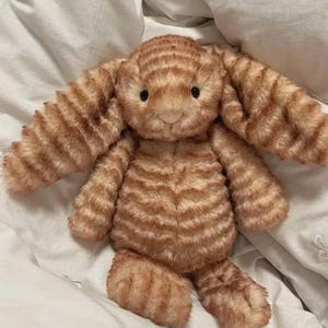 K&ouml;nnte beinhalten: Ein orange-braun gestreiftes Stofftier in Form eines Hasen. Das Kuscheltier hat lange Ohren, eine rosa Nase und schwarze Augen. Es liegt auf einem wei&szlig;en Bettlaken.