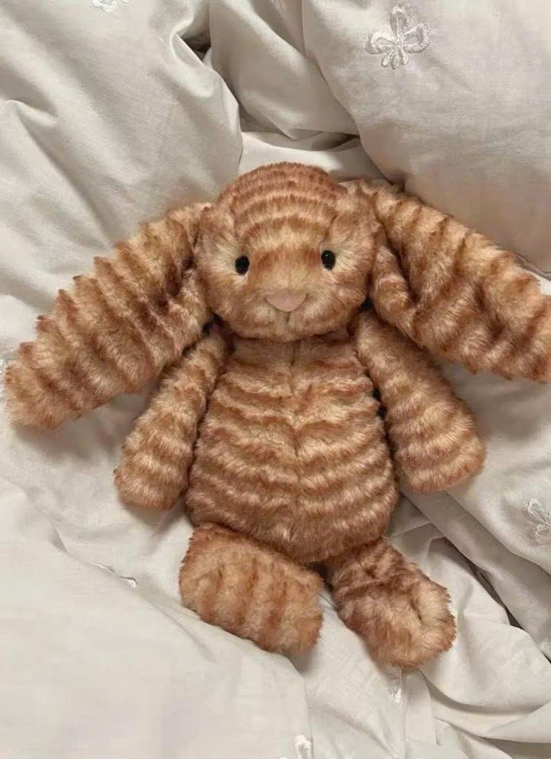 K&ouml;nnte beinhalten: Ein orange-braun gestreiftes Stofftier in Form eines Hasen. Das Kuscheltier hat lange Ohren, eine rosa Nase und schwarze Augen. Es liegt auf einem wei&szlig;en Bettlaken.
