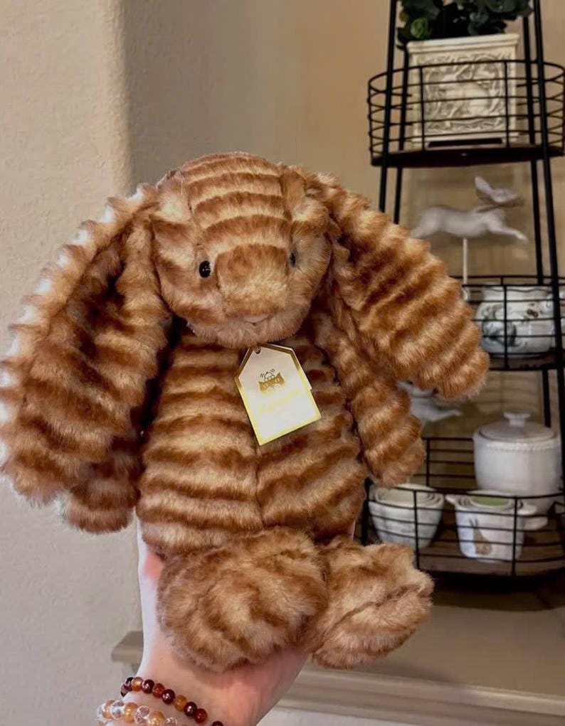 K&ouml;nnte beinhalten: Ein gestreiftes Stoffkaninchen in Braun- und Beiget&ouml;nen. Das Kuscheltier hat lange, schlaffe Ohren und ein kleines Etikett. Das Kaninchen ist weich und kuschelig, perfekt f&uuml;r eine Kinderspielzeugsammlung.