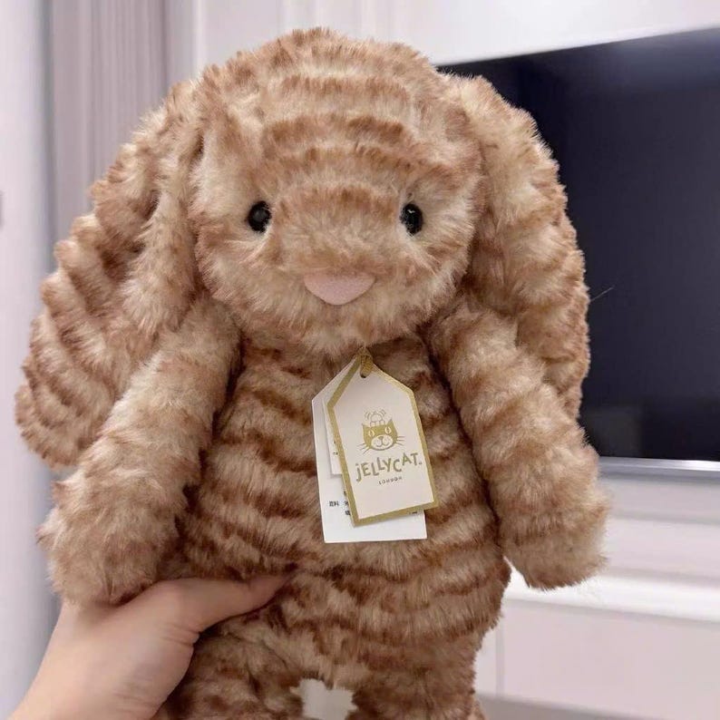 K&ouml;nnte beinhalten: Ein hellbraunes Pl&uuml;schkaninchen mit dunkelbraunen Streifen und langen, schlaffen Ohren. Das Stofftier hat eine rosa Nase und schwarze Augen. Ein Jellycat-Etikett ist angebracht.