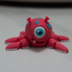 May include: A 3D-printed pink monster figurine with one large eye, blue spots, and multiple articulated arms. The monster has a smiling mouth with a single tooth and two small antennae.