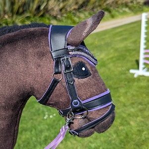 Puede incluir: Un caballo de juguete marrón con una melena negra y una brida negra y morada. El caballo de juguete tiene un mango de palo de madera y una cinta morada para las riendas. La imagen está tomada al aire libre en un césped.