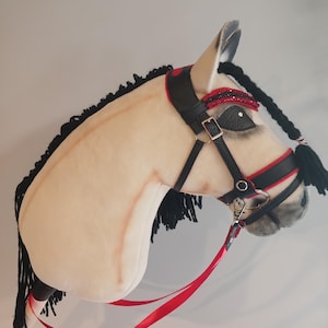 Puede incluir: Una cabeza de caballo de peluche crema con crin y cola negras. Cuenta con una brida negra y roja y una cinta roja para las riendas. El caballo tiene un ojo detallado y está diseñado para el juego imaginativo.