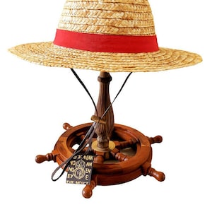 May include: A straw hat with a red band, displayed on a wooden ship's wheel base. The hat is made of woven straw and has a wide brim. The base is a dark brown colour and has a small tag with text.
