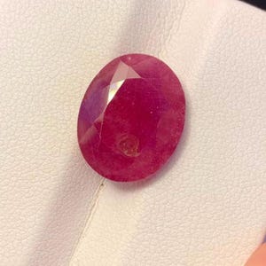 May include: A loose oval-cut gemstone with a deep magenta-red color and faceted surface. The stone displays internal light reflections and a small circular inclusion near the center, showcasing a translucent quality and a polished finish.