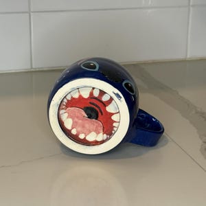 May include: A blue ceramic mug with a cartoonish design. The mug features a wide open mouth with red gums, white teeth, and a black tongue. The mug has two black eyes and a blue handle.