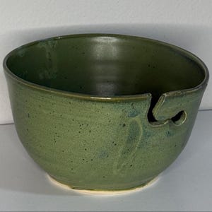 May include: A handmade, green ceramic yarn bowl with a unique design. The bowl features a curved opening to hold yarn and prevent tangling. The exterior has a speckled texture and a glossy finish, perfect for knitters and crocheters.
