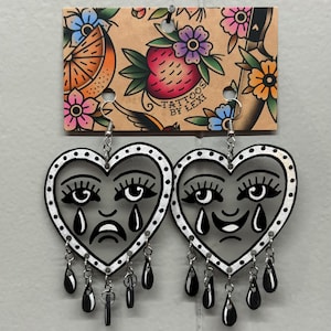 May include: A pair of heart-shaped earrings with a black and white design. Each earring features a face with a sad or happy expression, teardrops, and dangling black teardrop charms. The earrings hang from a decorative card with floral and fruit illustrations and the text "TATTOOS BY LEXI."