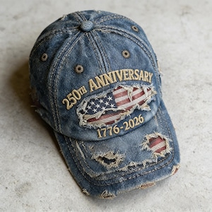 Puede incluir: Gorra de béisbol de mezclilla desgastada con texto bordado "250th Anniversary 1776-2026" y un diseño de bandera estadounidense rasgada. La gorra presenta un aspecto desgastado con desgaste visible y una visera curva.