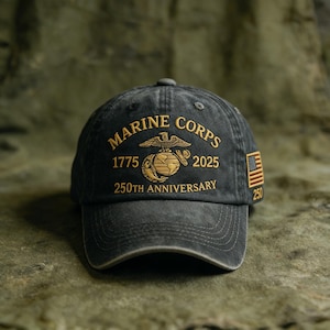 May include: A distressed black baseball cap with gold embroidered text and graphics. The text reads "MARINE CORPS 1775 2025 250TH ANNIVERSARY" with an eagle, globe, and anchor emblem. An American flag patch is on the side.