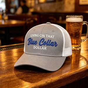 Puede incluir: Gorra de camionero gris con parte trasera de malla blanca y visera curva. La gorra presenta el texto bordado "LIVING ON THAT Blue Collar DOLLAR" en azul y blanco. Un vaso de cerveza se encuentra en el fondo.