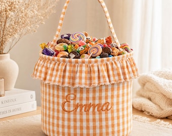 May include: A cylindrical fabric basket featuring an orange and white gingham pattern with a ruffled top edge and a single handle. The name Emma is embroidered in brown script on the front. The basket is filled with a variety of colorful candies.