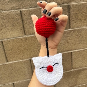May include: A handmade crochet keychain. It features a red ball and a white cat face with black stitched eyes and a red nose. The ball is connected to the cat face with a black cord.