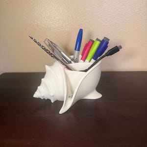 May include: A white, seashell-shaped desk organizer filled with pens, markers, and a USB cable. The organizer is made of ceramic or a similar material and sits on a dark brown surface. The pens and markers are various colors, including blue, pink, and green.