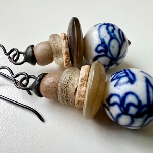 May include: A pair of earrings with a blue and white porcelain bead and a stack of brown, beige, and white beads. The earrings have a black wire hook.