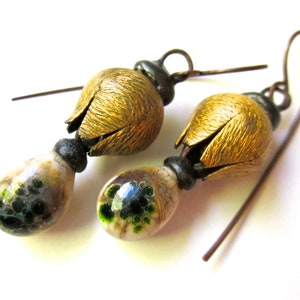 May include: A pair of drop earrings featuring textured, golden-toned metal flower caps attached to dark metal hooks. Below each flower cap is a smooth, teardrop-shaped bead with a cream-colored base and mottled green and black speckled patterns.