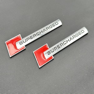 May include: Two silver "SUPERCHARGED" car emblems with a red square design. The rectangular emblems have rounded ends and are set against a dark grey textured background. The text is in a sans-serif font.