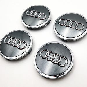 May include: Four gray and silver Audi wheel center caps. Each cap features the Audi logo in silver against a gray background, with a silver trim. The caps are circular and appear to be new.