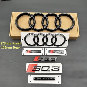 May include: A collection of black car emblems, including the Audi rings, S Line, and SQ3 badges. The front emblem measures 27.3cm, and the rear emblem is 19.2cm. The emblems are displayed on a white backing and a cardboard box.