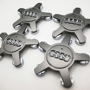May include: Four silver-gray Audi wheel center caps. Each cap features the Audi logo and a five-spoke design with chrome accents. The caps are arranged on a white surface, showcasing their metallic finish.