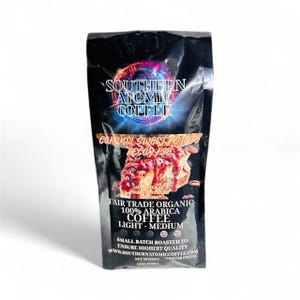 Op de afbeelding: Een zwarte zak Southern Atomic Coffee. De zak heeft het logo van het merk en de woorden "Caramel Sweet Potato Pecan Pie". Het is Fair Trade Organic, 100% Arabica, Light-Medium roast. Het nettogewicht is 340g.