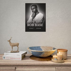 May include: A black and white poster featuring a portrait of a man with the text LONG LIVE ROB BASE 1967 - 2026. The poster is displayed on a wall above a wooden surface containing a wooden deer figurine, books, a woven bowl, and candles.