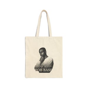 May include: Cream canvas tote bag with a black and white portrait of a man. The text "LONG LIVE ROB BASE" is printed beneath the image. The bag has two handles and is made of a canvas material.
