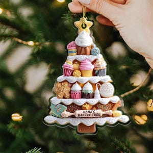 May include: A Christmas tree ornament shaped like a bakery tree, featuring cupcakes, cookies, and other baked goods. The ornament has a gold star at the top and the text "NANA'S BAKERY TREE" on a banner. The ornament is held by a hand.