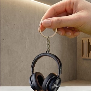 May include: A black headphone keychain with the word "Novice" printed on the side. The keychain is held by a silver chain and a silver key ring. The headphones have a padded headband and earcups.