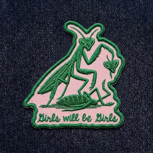 May include: Embroidered patch featuring a green praying mantis design on a pink background, outlined in green. The text "Girls will be Girls" is at the bottom. The patch is set against a dark blue denim background.