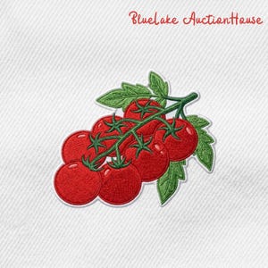 May include: An embroidered patch featuring a cluster of bright red tomatoes on a vine with green leaves, set against a white fabric background. The text Bluelake AuctionHouse is printed in red script at the top of the white fabric surface.