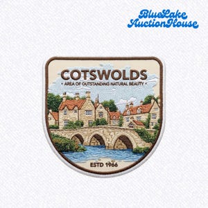 May include: A shield-shaped embroidered patch featuring a landscape of stone cottages and a stone bridge over water. The text reads COTSWOLDS AREA OF OUTSTANDING NATURAL BEAUTY and ESTD 1966. The patch has a brown border and a cream-colored background.