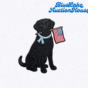 May include: An embroidered patch featuring a black Labrador retriever wearing a light blue ribbon collar and holding a small American flag in its mouth. The text BlueLake AuctionHouse appears in blue script in the upper right corner.