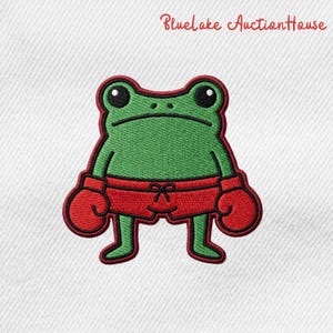 May include: Embroidered patch featuring a green frog wearing red boxing gloves and shorts. The frog has a serious expression and is outlined in red. The text "BlueLake AuctionHouse" is in red script at the top.