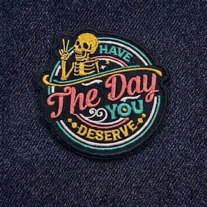 May include: Embroidered patch with a skeleton giving a peace sign, with the text "Have The Day You Deserve". The patch has a black background with a colorful design, including yellow, pink, and teal.